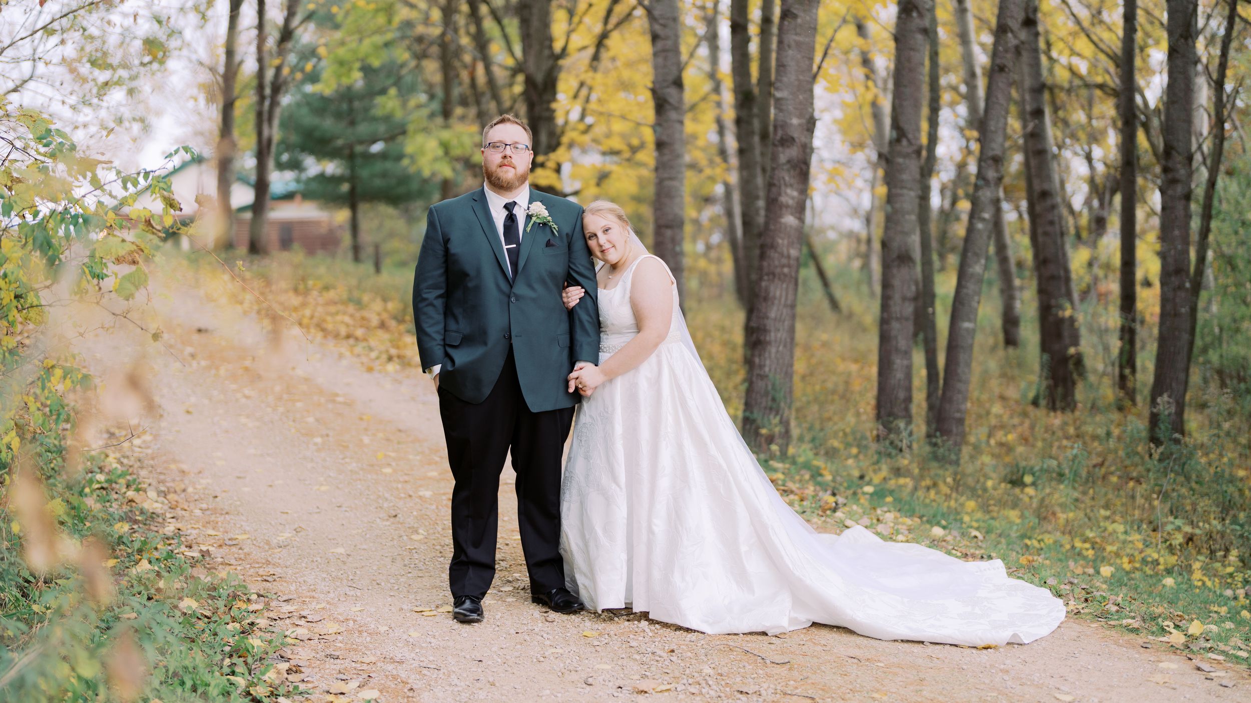 A couple in wedding attire walks along a winding autumn forest path surrounded by golden fall foliage.