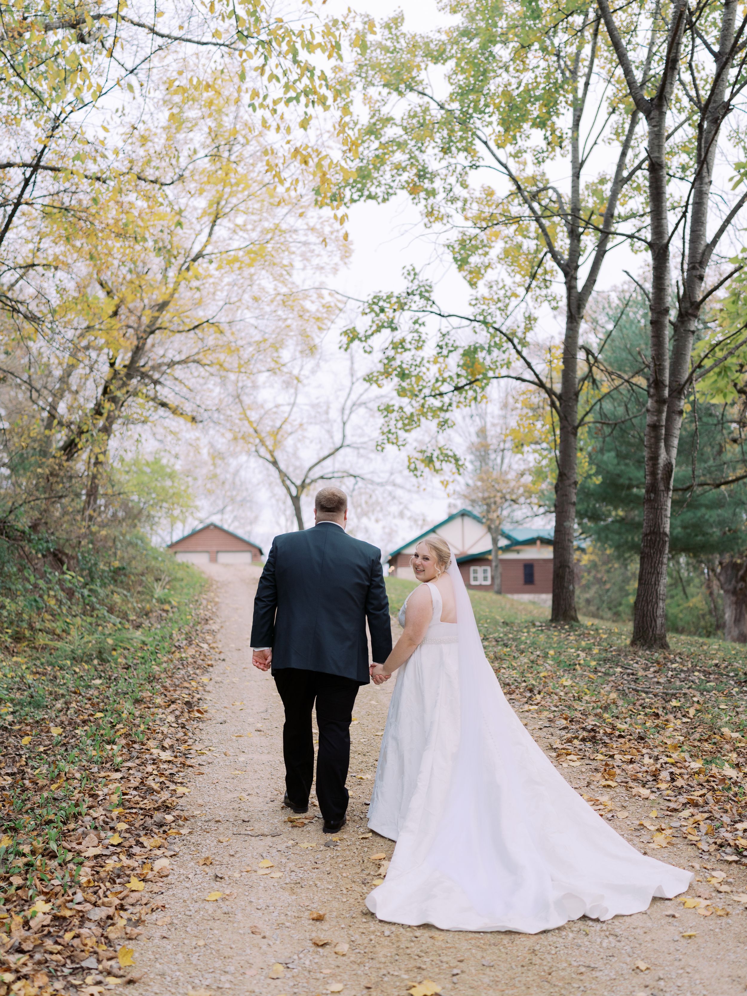 A couple in wedding attire walks down a leaf-covered path through autumn trees on their special day.