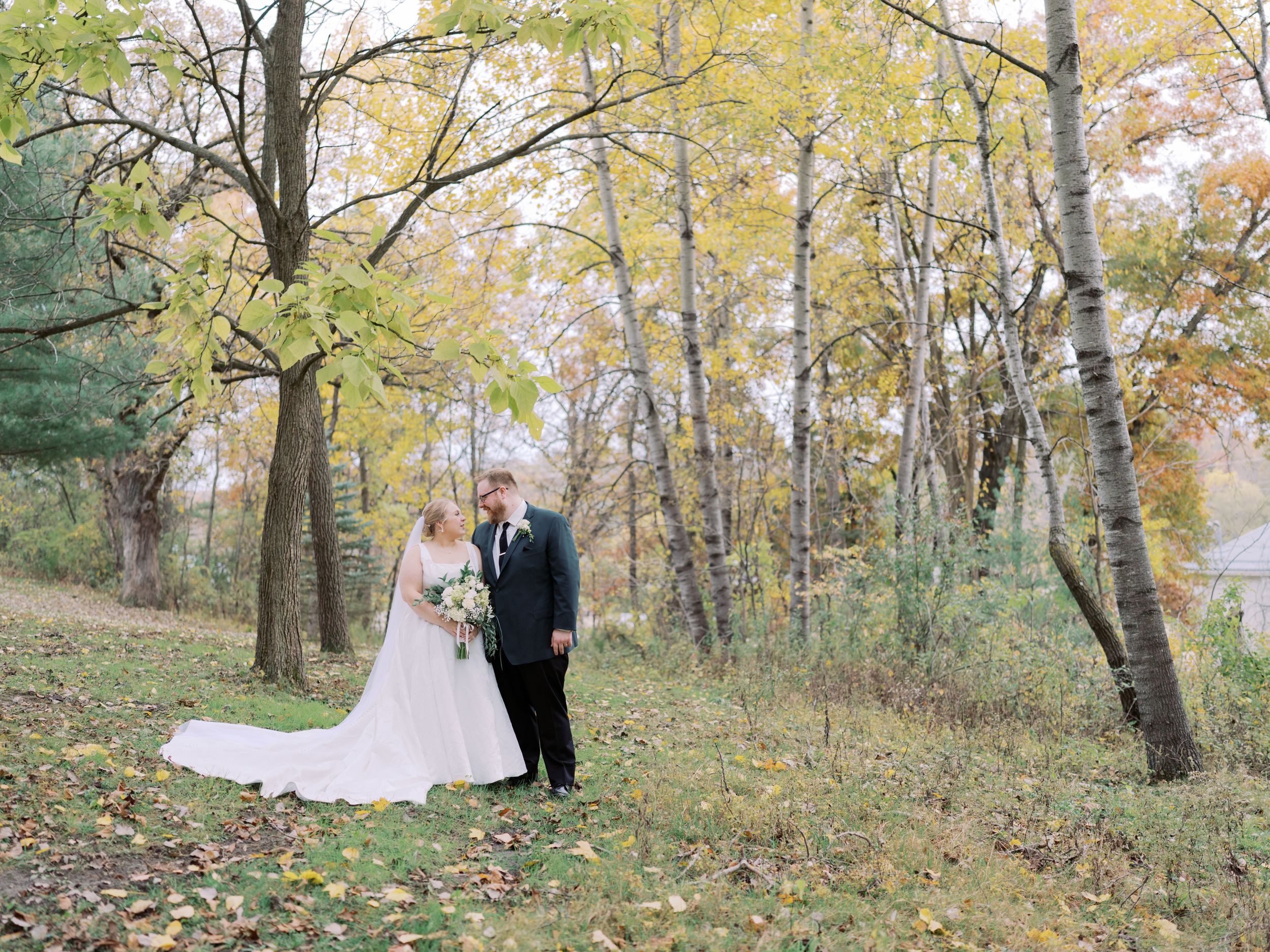 A romantic wedding couple poses in a serene autumn forest setting with golden yellow leaves and tall trees.