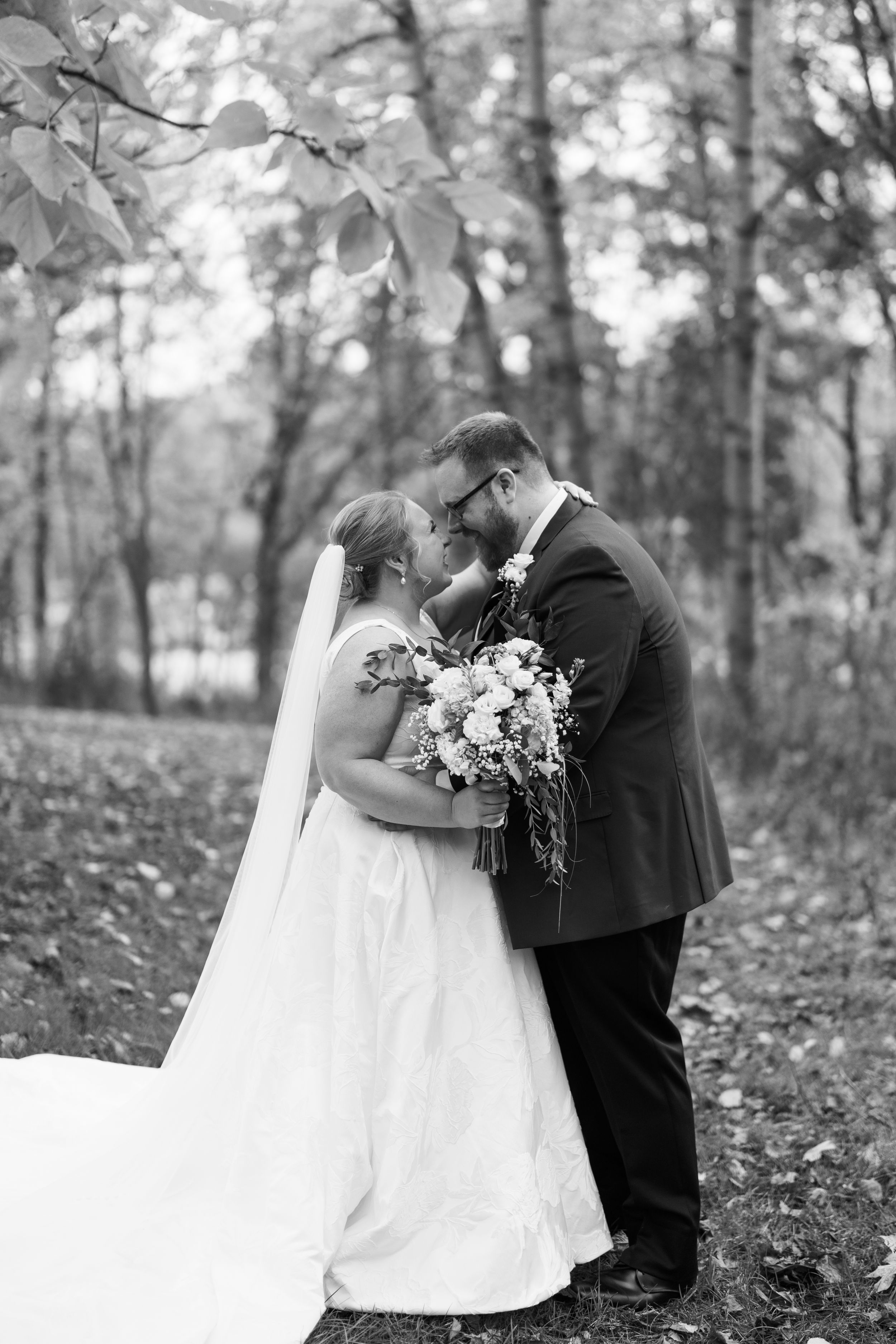A romantic black and white wedding photo of a couple sharing an intimate moment in a wooded outdoor setting.