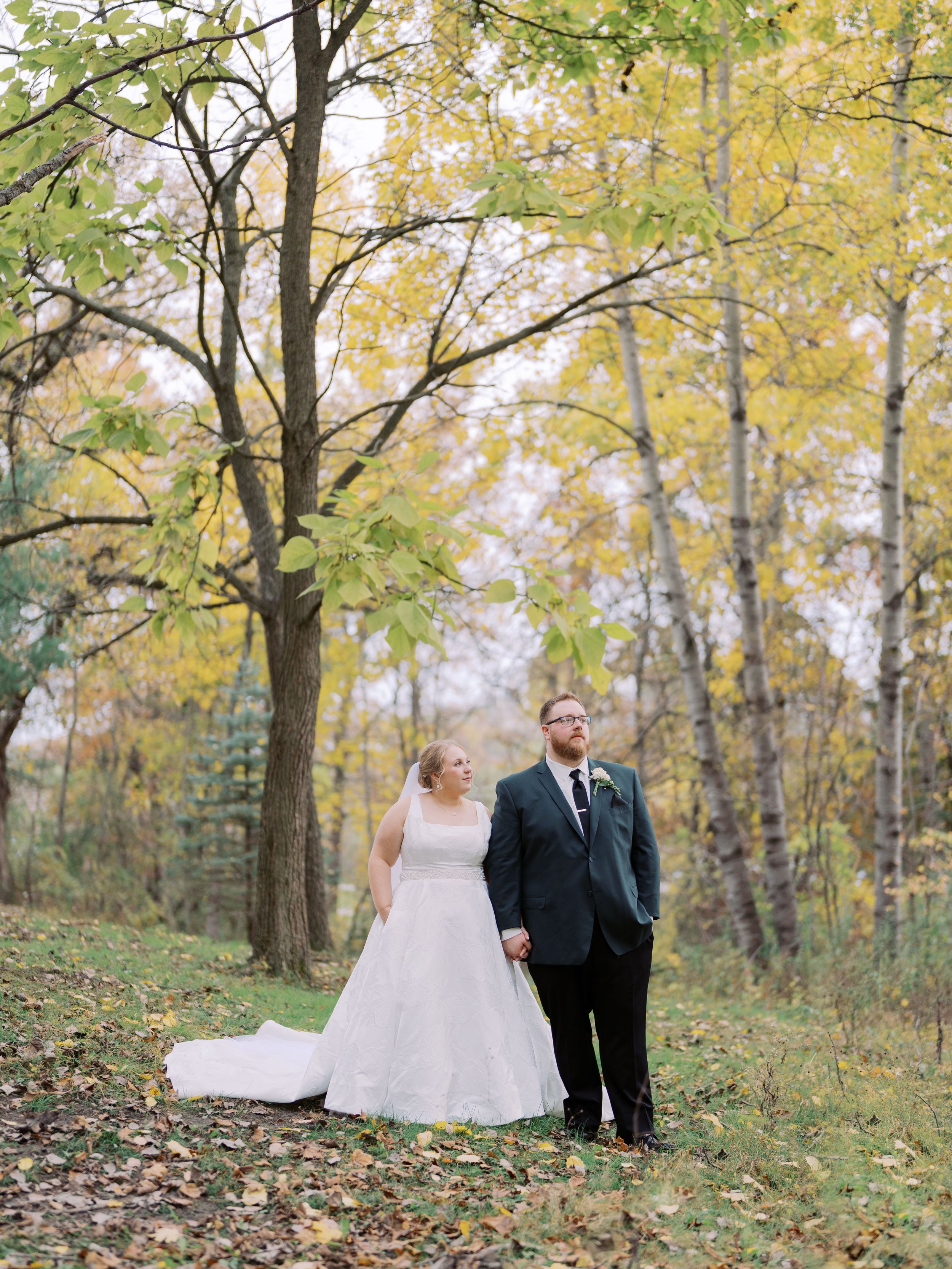 A couple poses together in an autumn forest setting with yellow leaves and trees surrounding them.