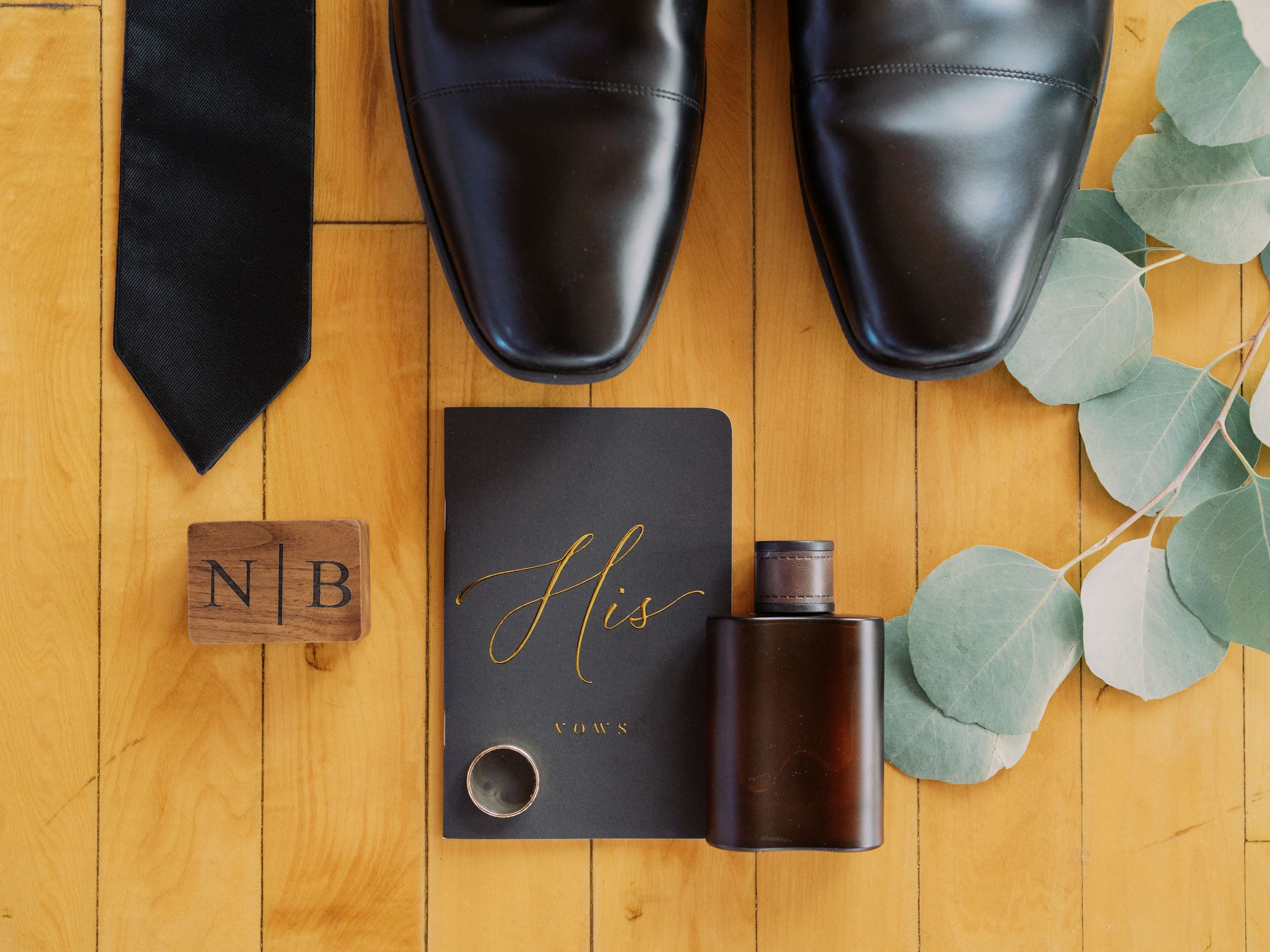 Black leather shoes and accessories arranged on wooden background with eucalyptus leaves and wedding details.