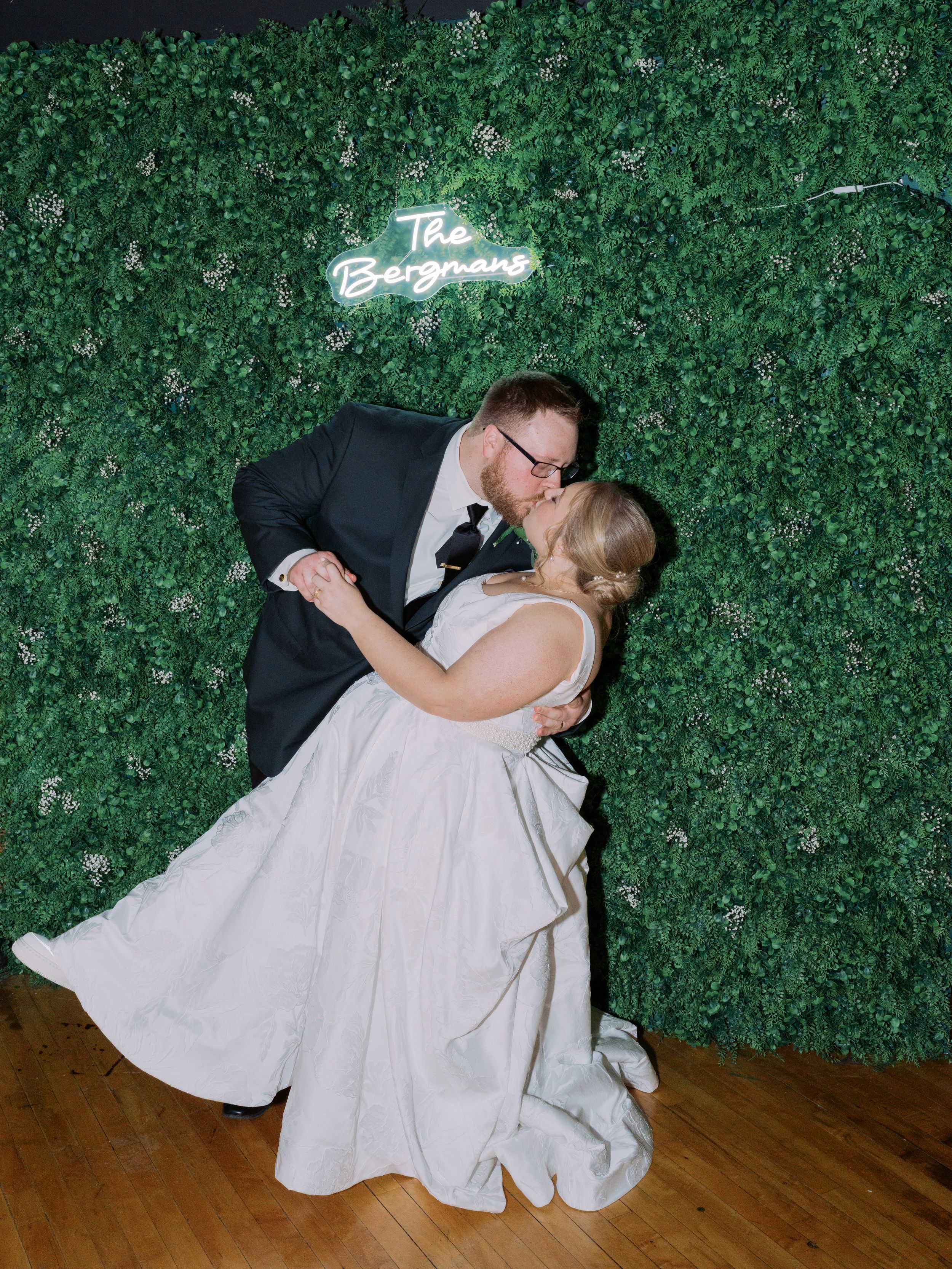 A couple in wedding attire dances together on a wooden floor against a lush green foliage wall backdrop.