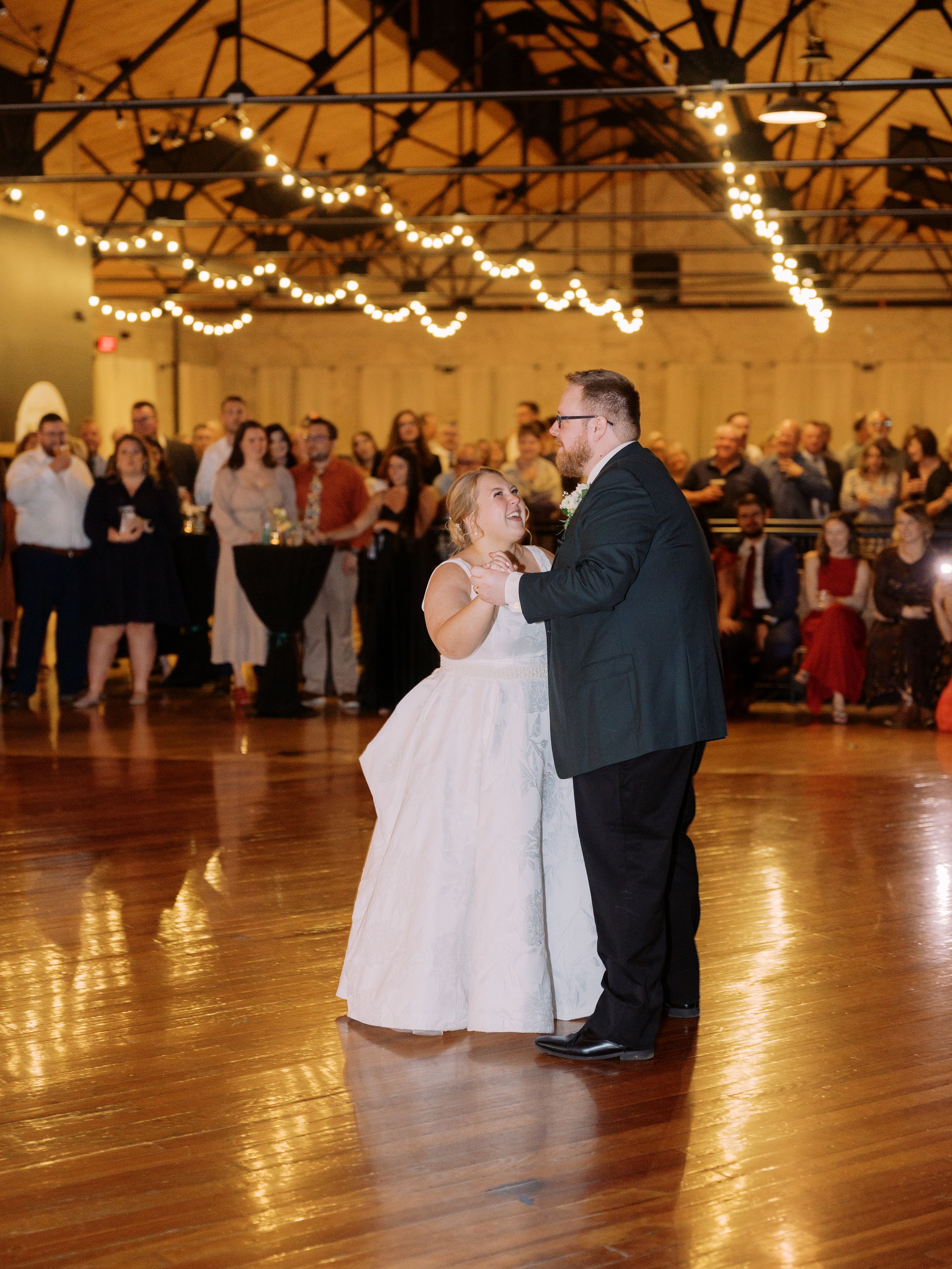 Wedding couples dancing in a large wooden-floored venue decorated with string lights overhead.