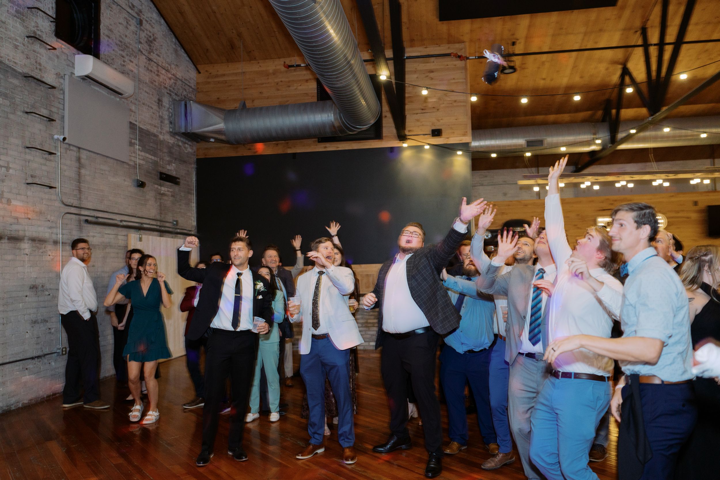 Wedding guests dance and celebrate on a rustic wooden dance floor under warm lighting at an indoor reception venue.