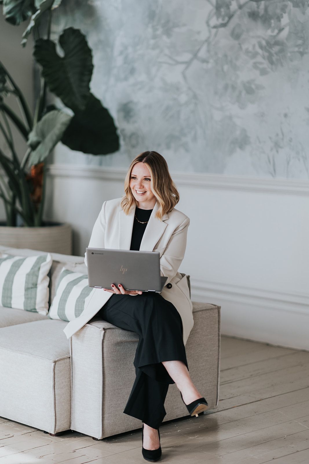 Professional lifestyle photo series showing someone in a black dress and beige blazer sitting on a gray couch in different poses.