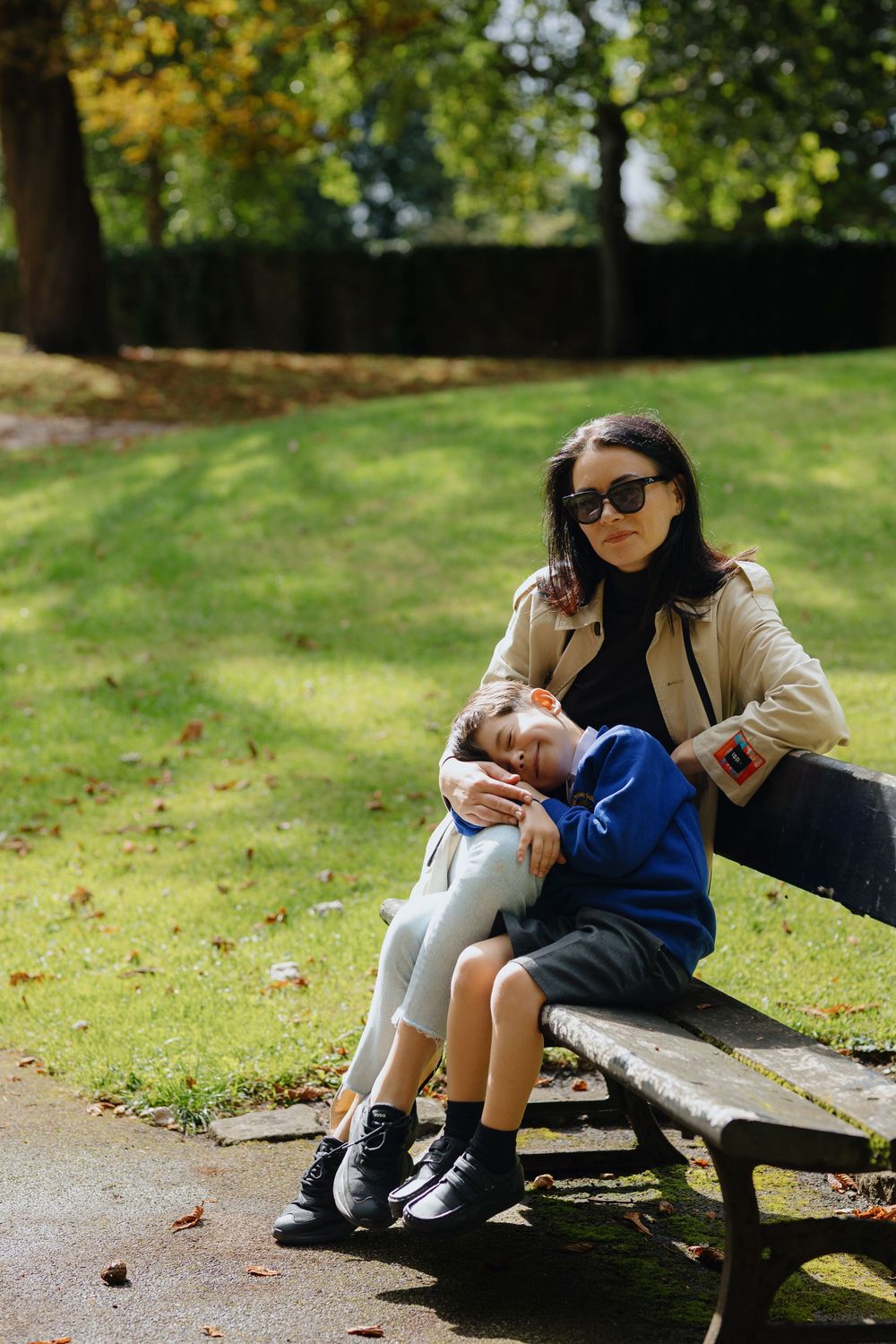 Person sitting on bench in autumn park using mobile phone while resting on grassy lawn in sunlight.
