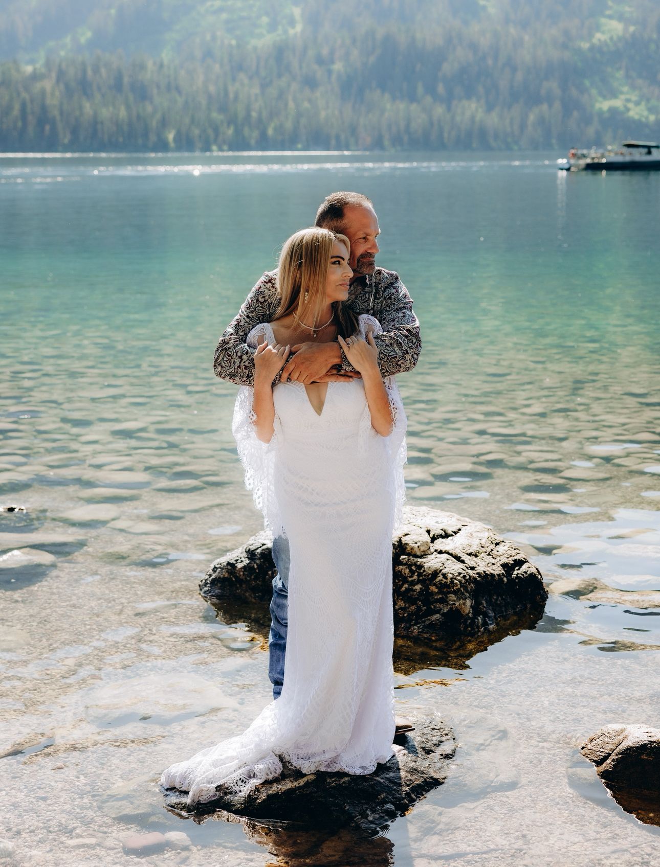 A romantic couple shares tender moments by a serene mountain lake with evergreen forest and calm waters in the background.
