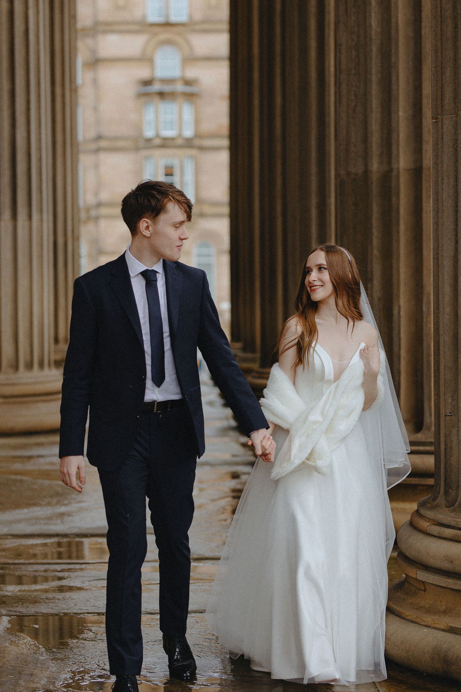 A couple in wedding attire poses playfully between stone columns of a historic building.