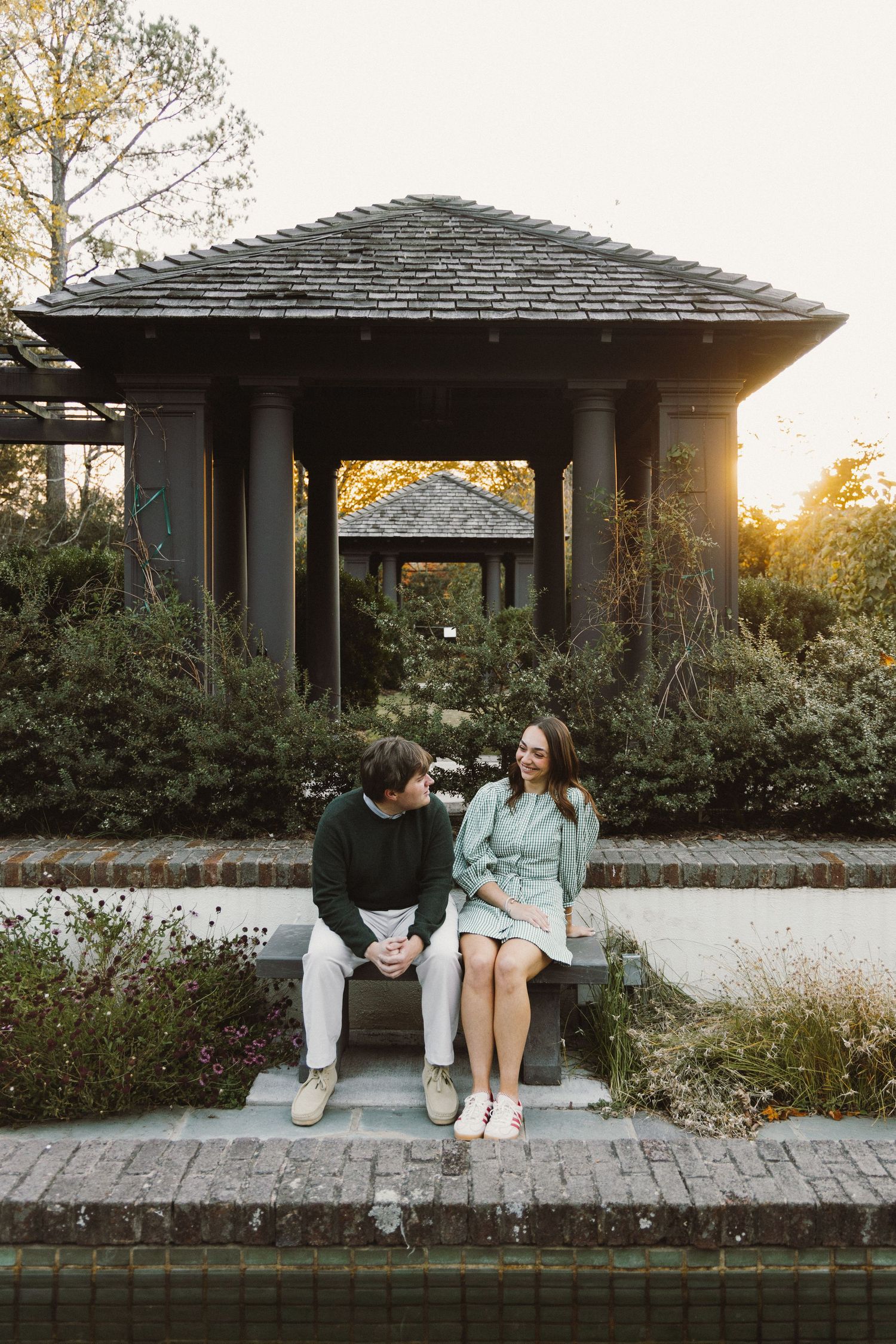 Two people sitting together at sunset under a wooden gazebo structure in a garden setting.