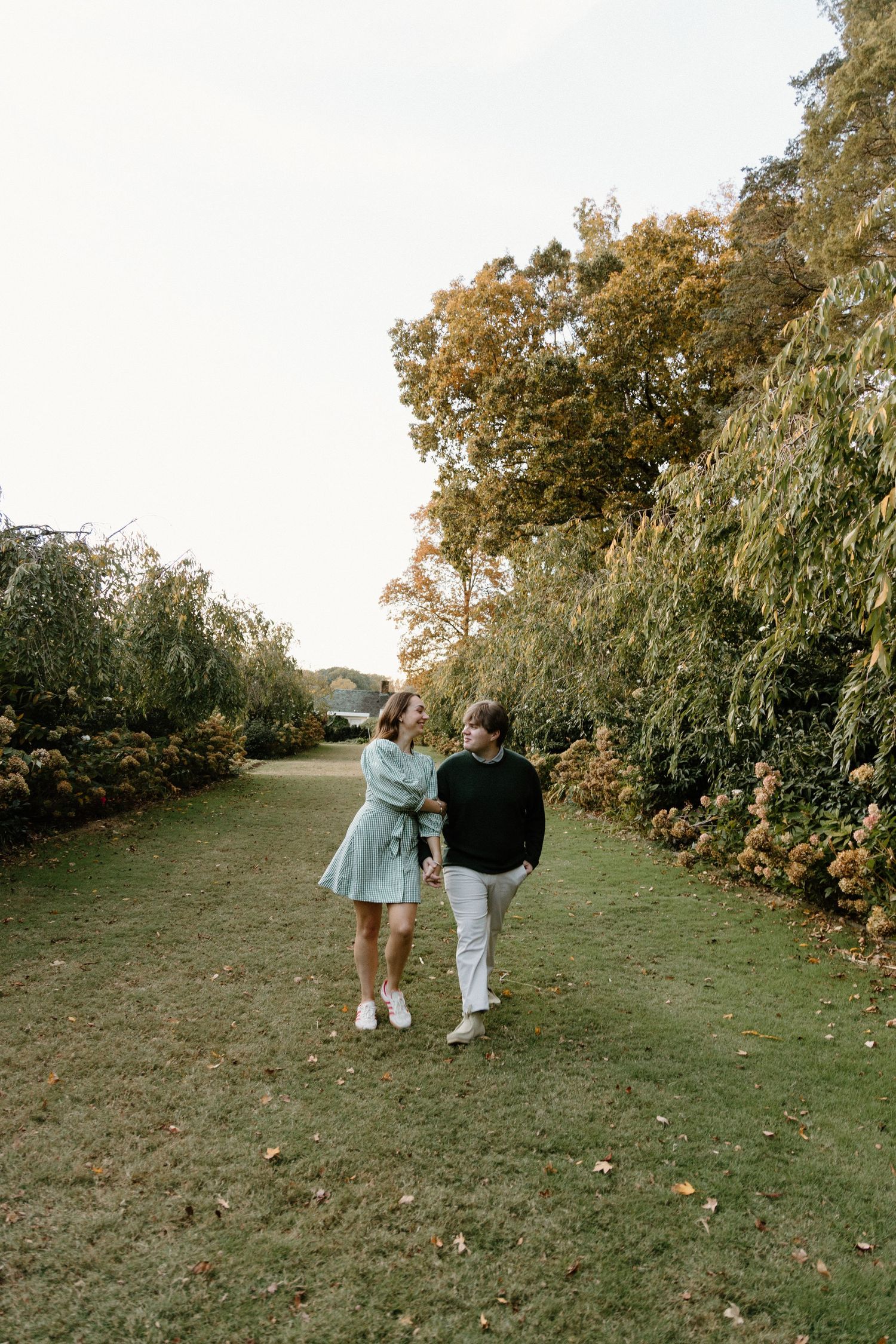 A couple walks hand in hand down a garden path lined with autumn trees and flowers on a peaceful afternoon.