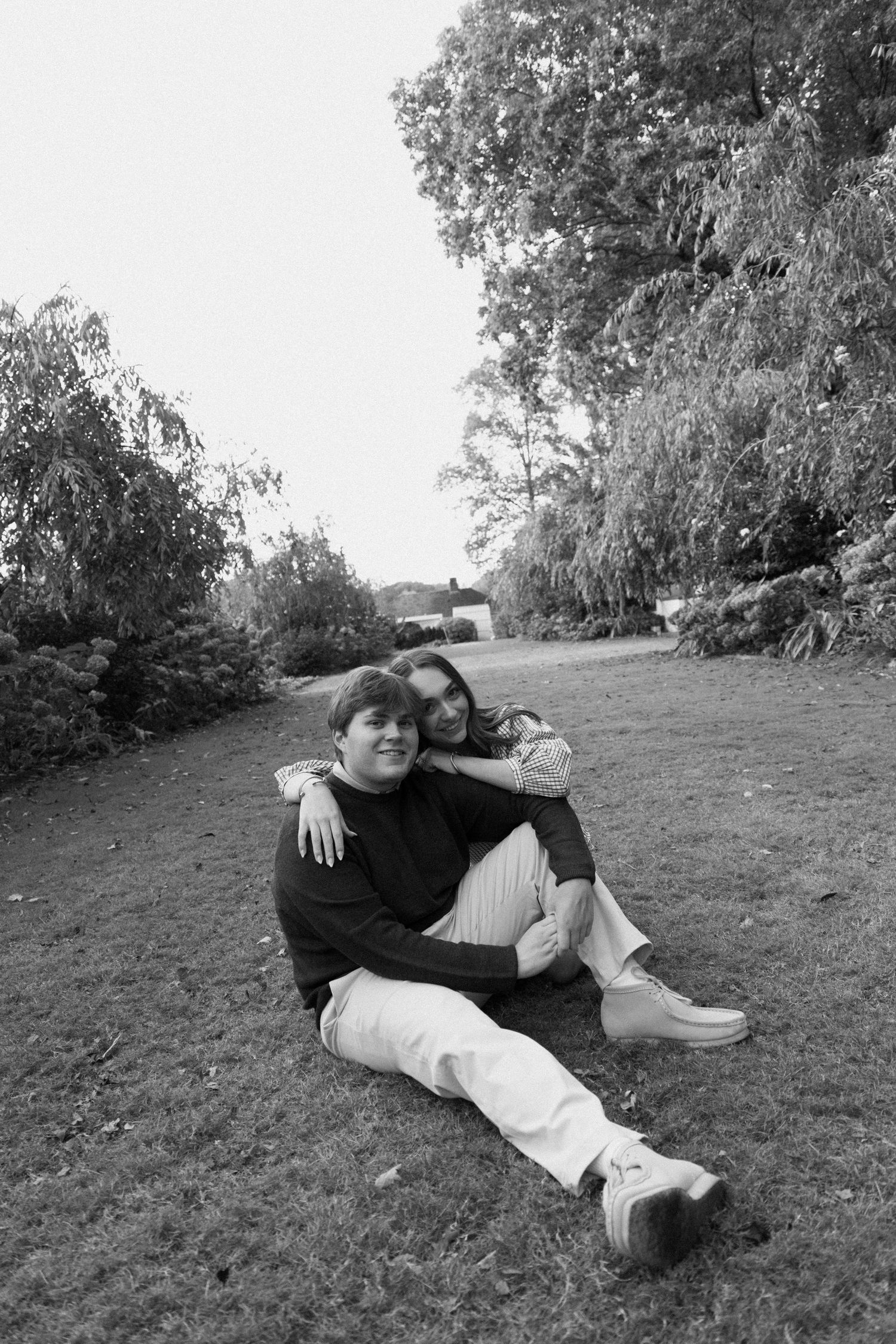 A couple relaxes together on grass beneath tall trees in a black and white photograph.