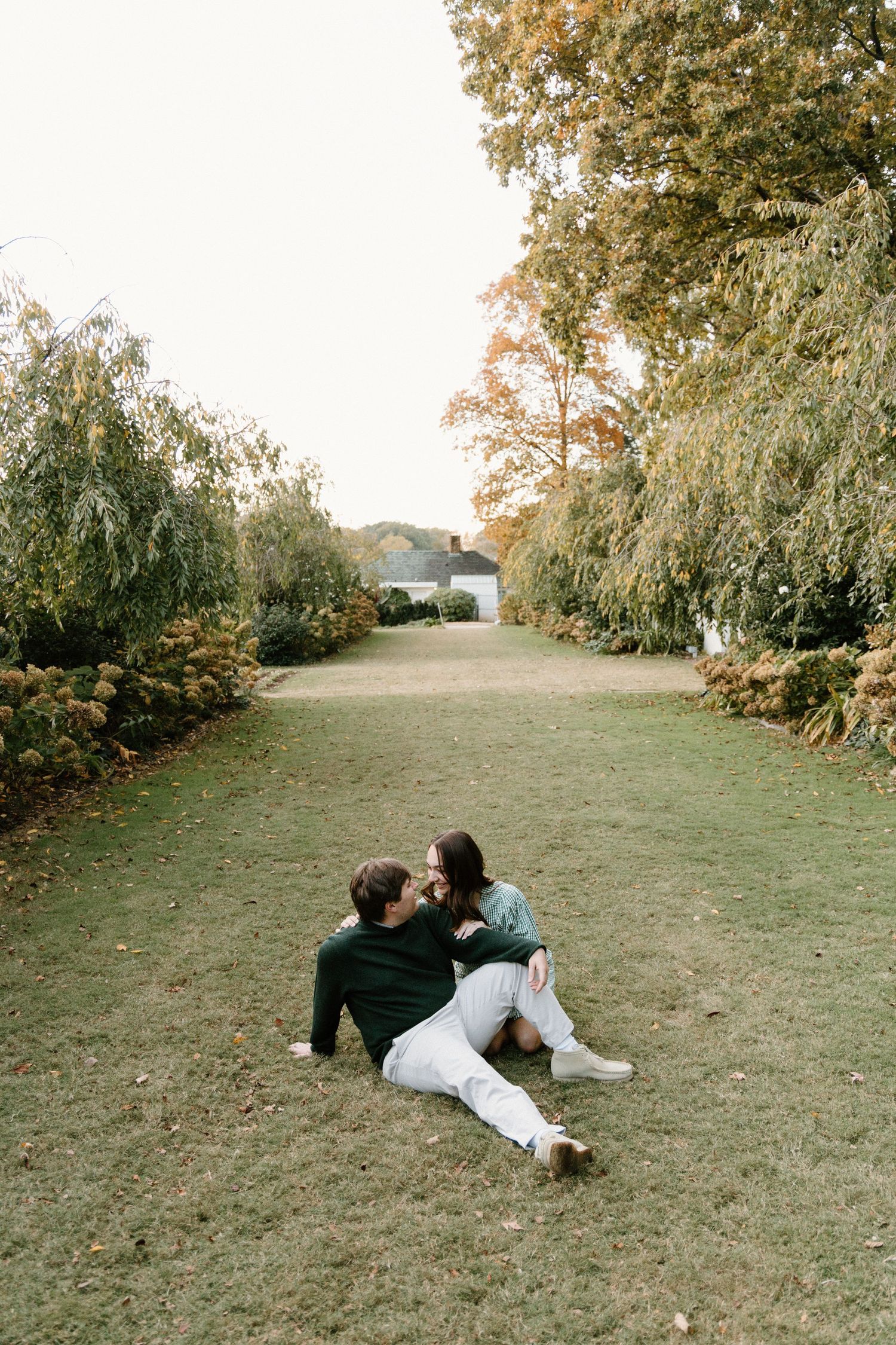 A couple lies together on a grassy hillside surrounded by trees during autumn.