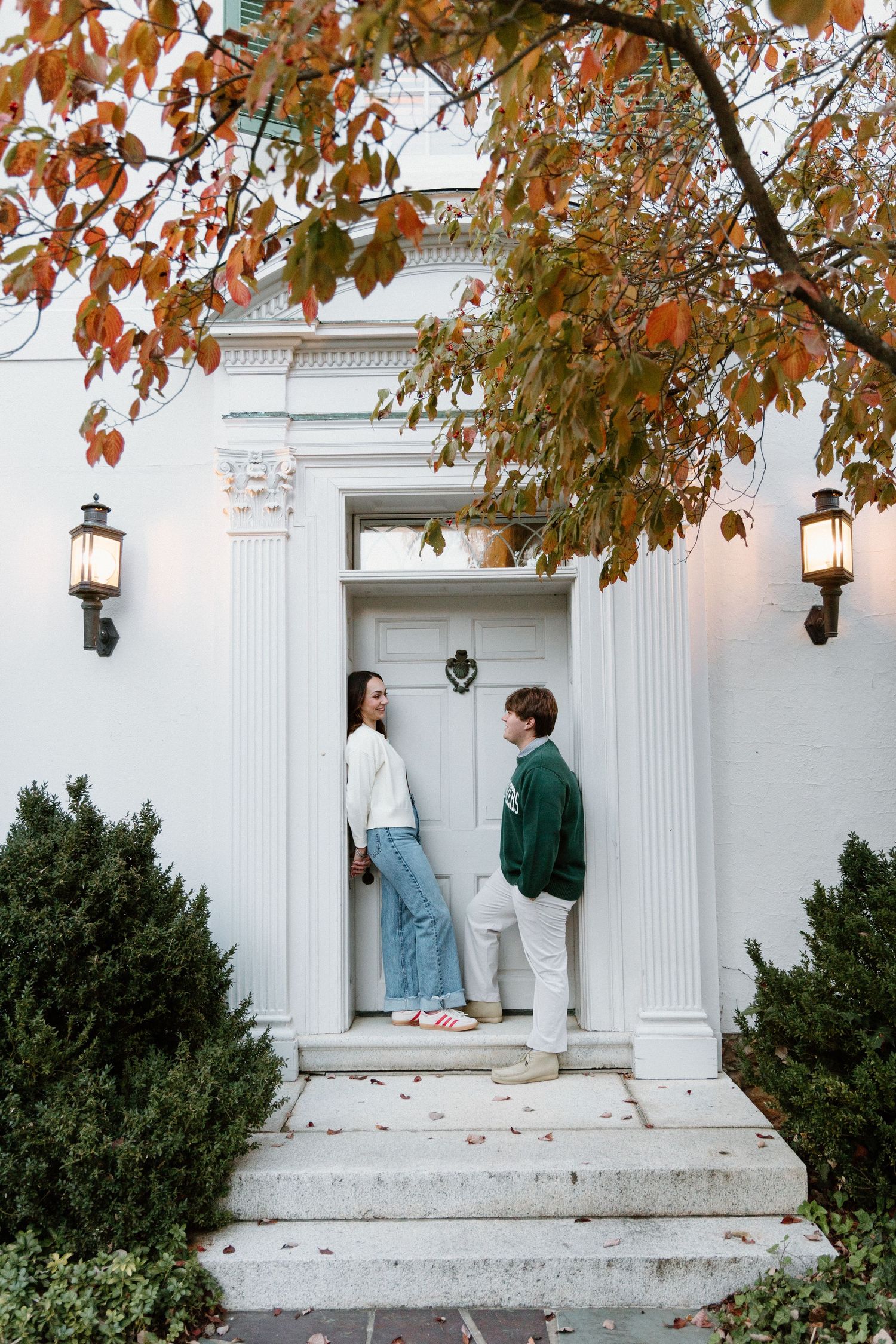 A couple stands on white stone steps beneath autumn leaves in a romantic outdoor engagement photo series.