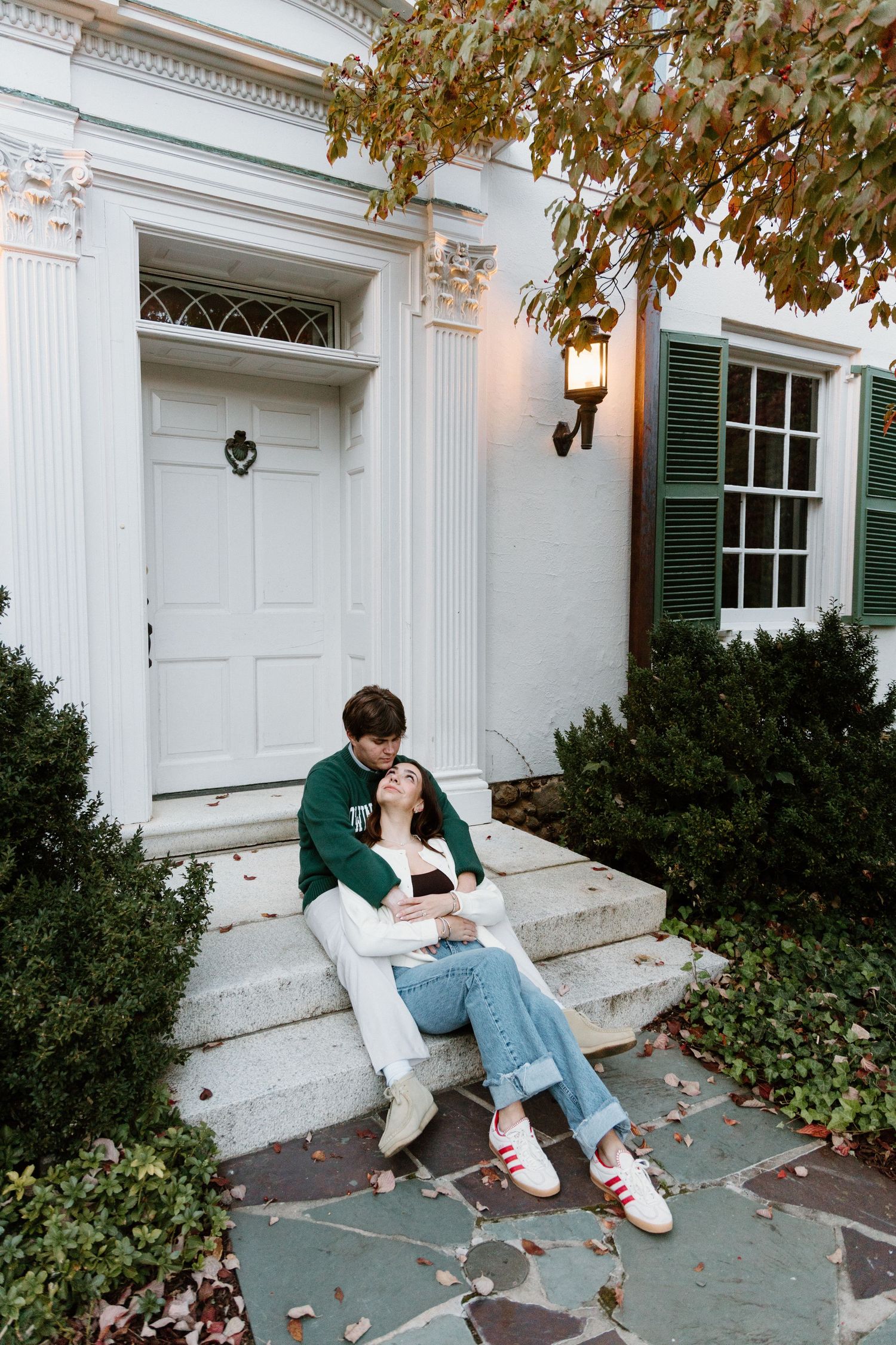 A couple sits together on white steps of a classic colonial style house with green shutters and autumn leaves falling.