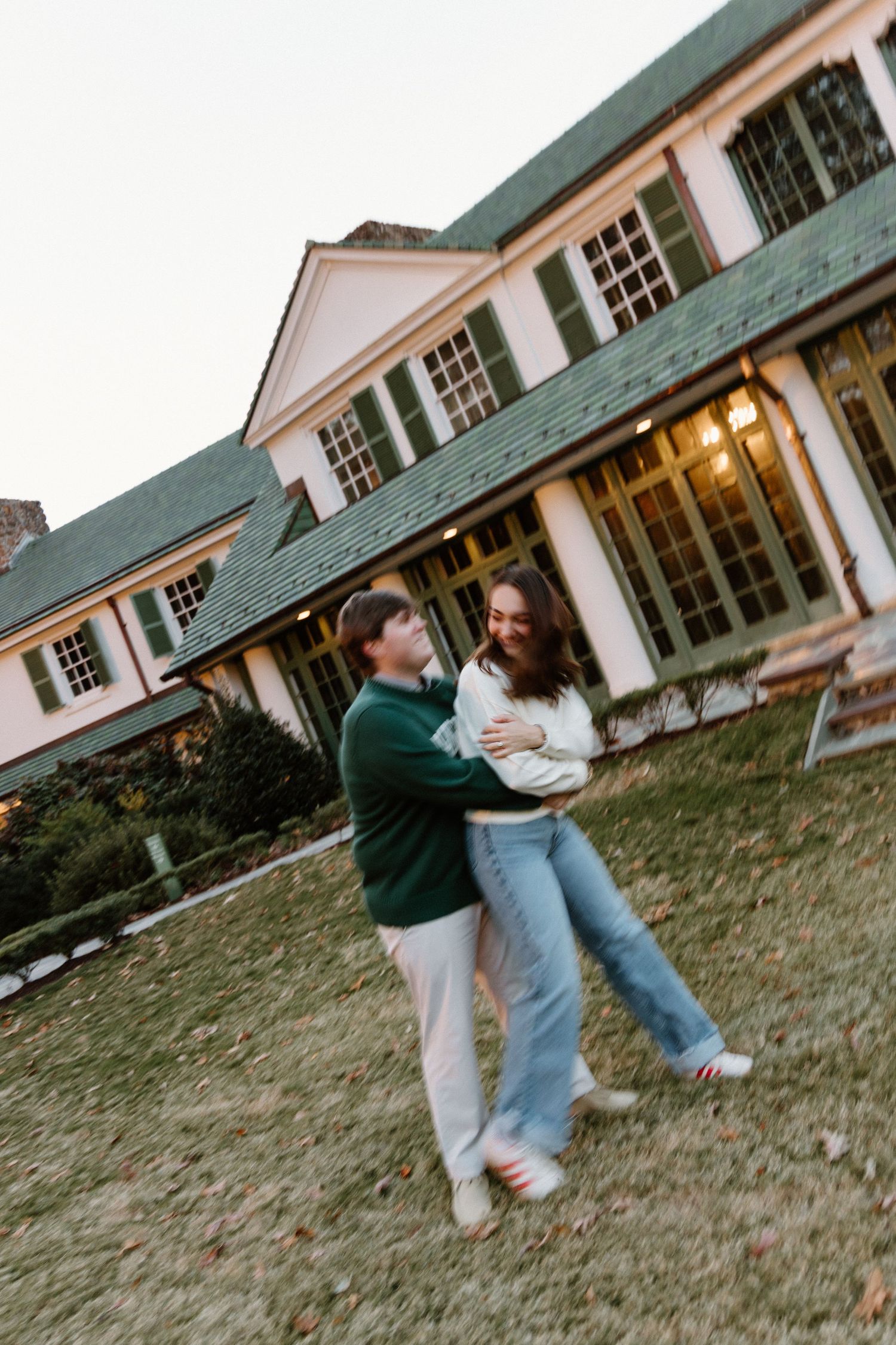 A couple dances and spins together on a grassy lawn in front of a white colonial-style building at dusk.