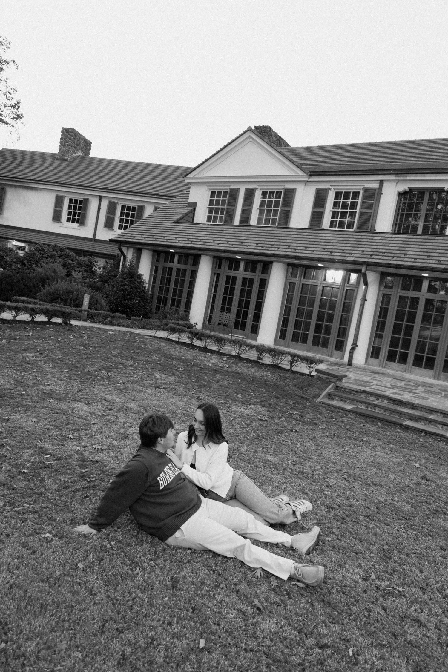 A couple lies on the grass in front of a large white colonial-style building with many windows.