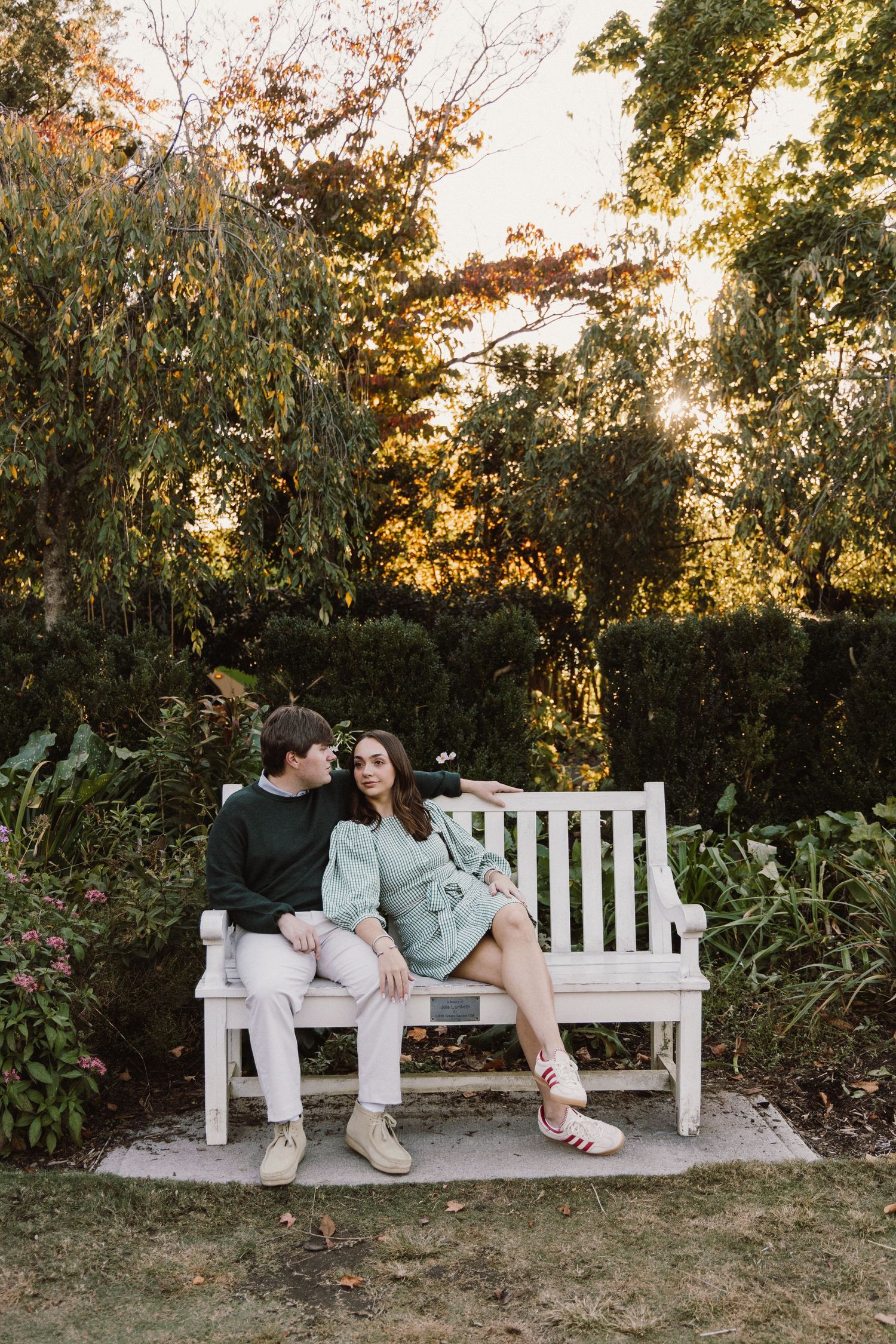 A couple sits together on a white garden bench surrounded by lush greenery during golden hour sunset.