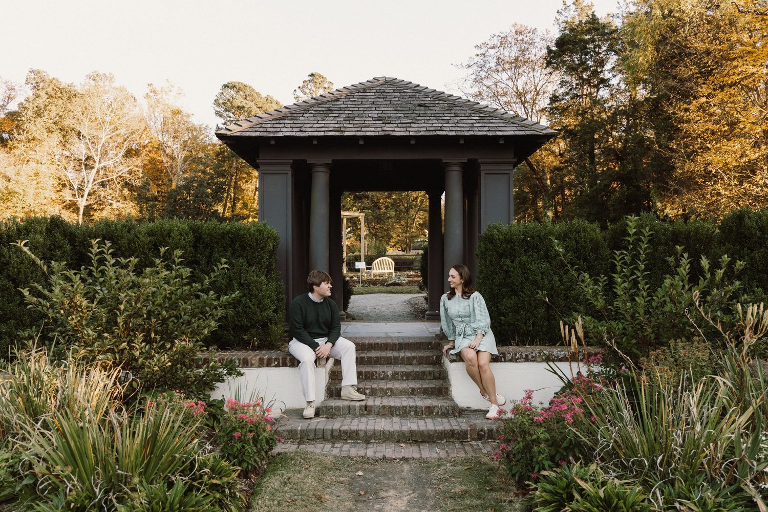 A romantic couple stands in front of a rustic gazebo surrounded by lush autumn gardens at sunset.