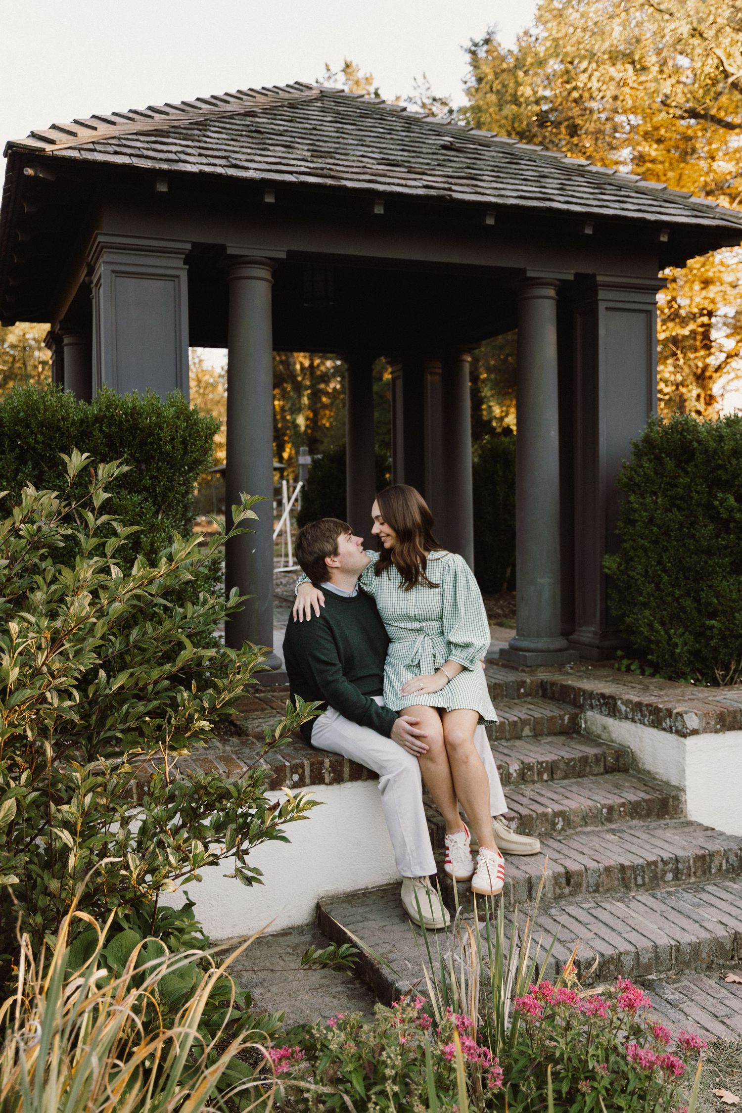 A couple sits together on stone steps of a covered porch surrounded by autumn foliage and pink flowers.