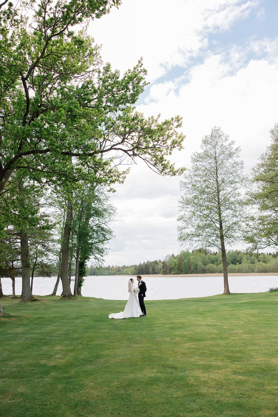 A couple sits on a grassy lawn overlooking a serene lake with tall trees framing the peaceful waterfront scene.