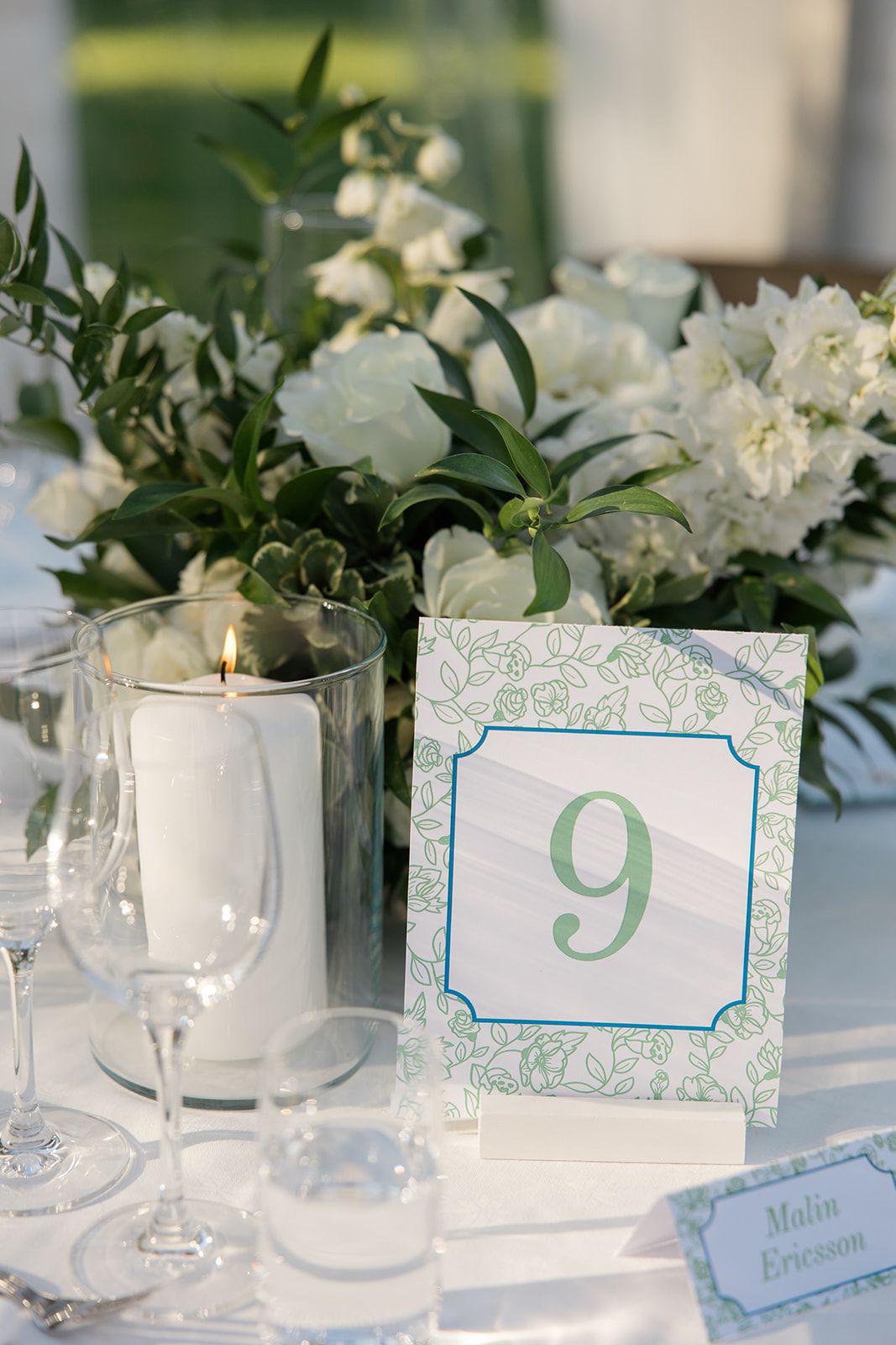 Elegant white wedding reception table setting with floral centerpieces, table numbers, and glowing candles in glass holders.