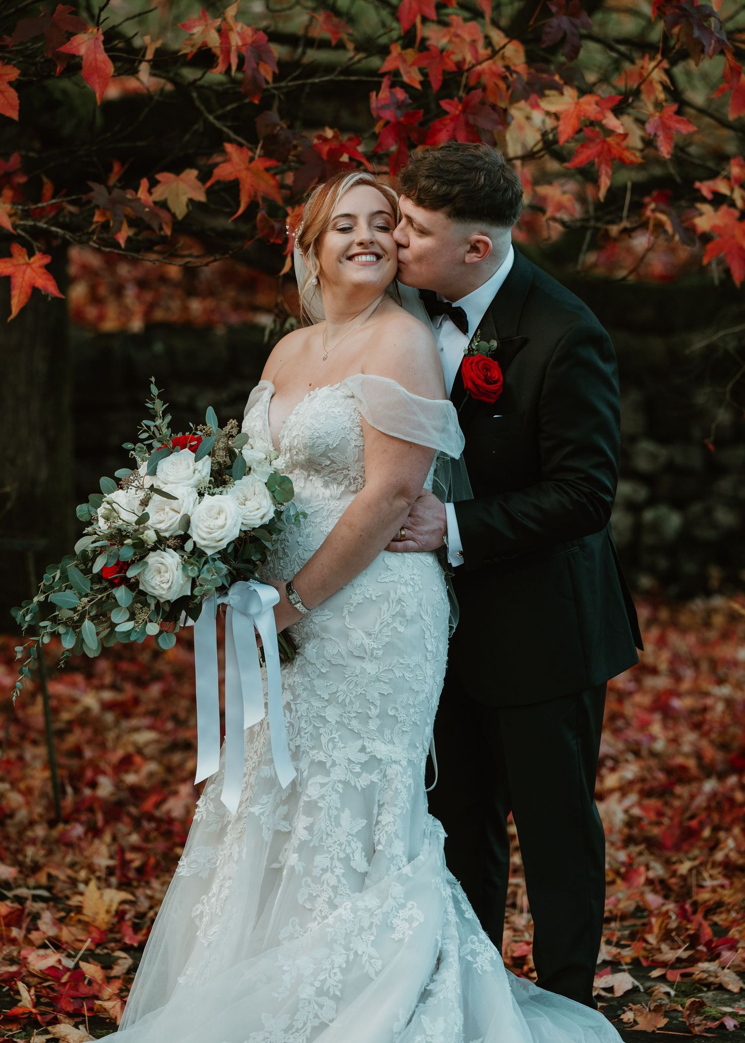 Romantic autumn wedding couple embraces under vibrant red maple leaves during outdoor photo session.