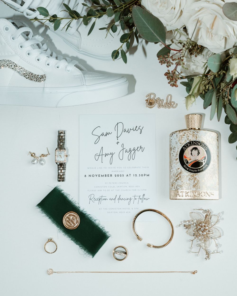 Wedding details flatlay with rings, invitations, perfume bottles and eucalyptus leaves on white marble background.