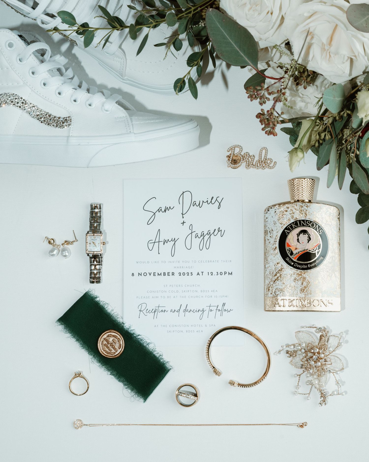 Wedding details flatlay with rings, invitations, perfume bottles and eucalyptus leaves on white marble background.