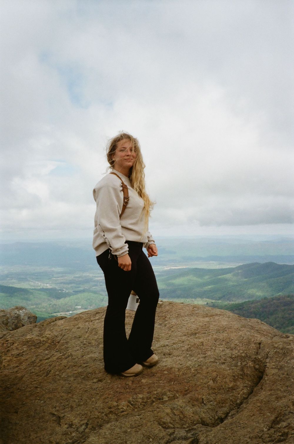 Person in white sweater and dark pants sits contemplatively on rocky mountain peak overlooking vast green valley landscape.
