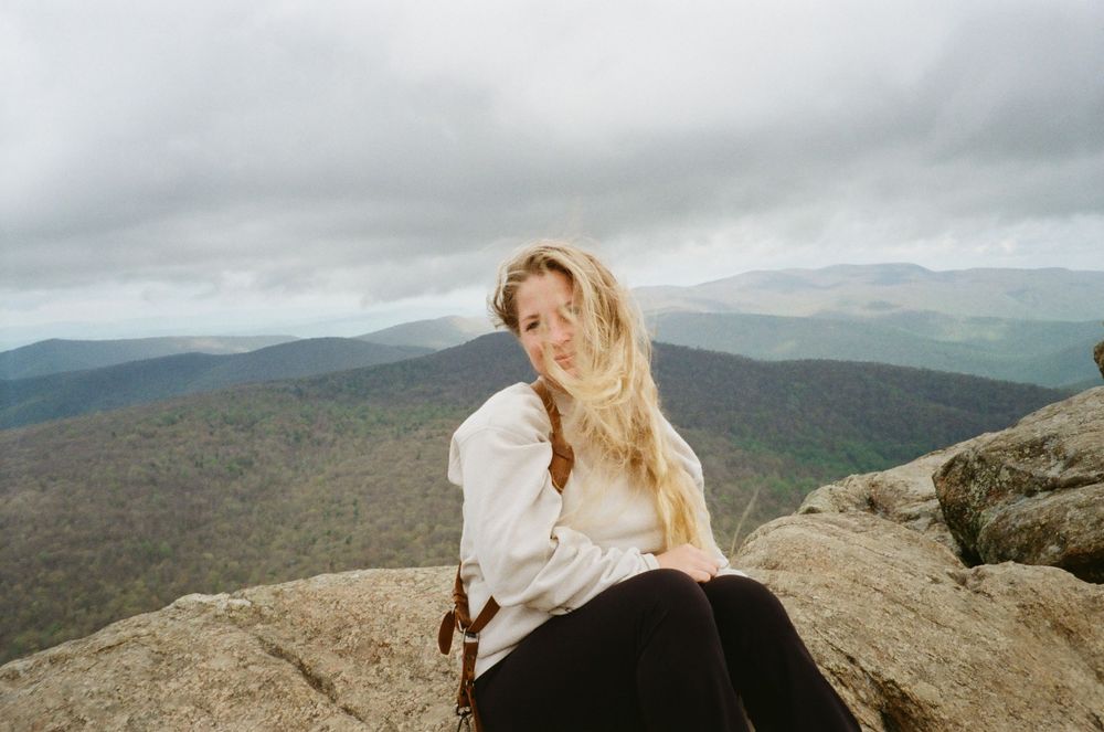 A hiker stands on a rocky mountain summit overlooking a vast landscape of rolling hills and valleys on a cloudy day.