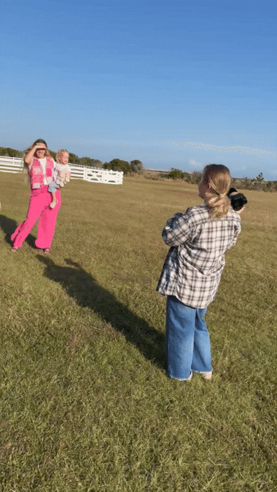 A person in a plaid shirt photographs pink baby clothes hanging on a clothesline in a grassy field.