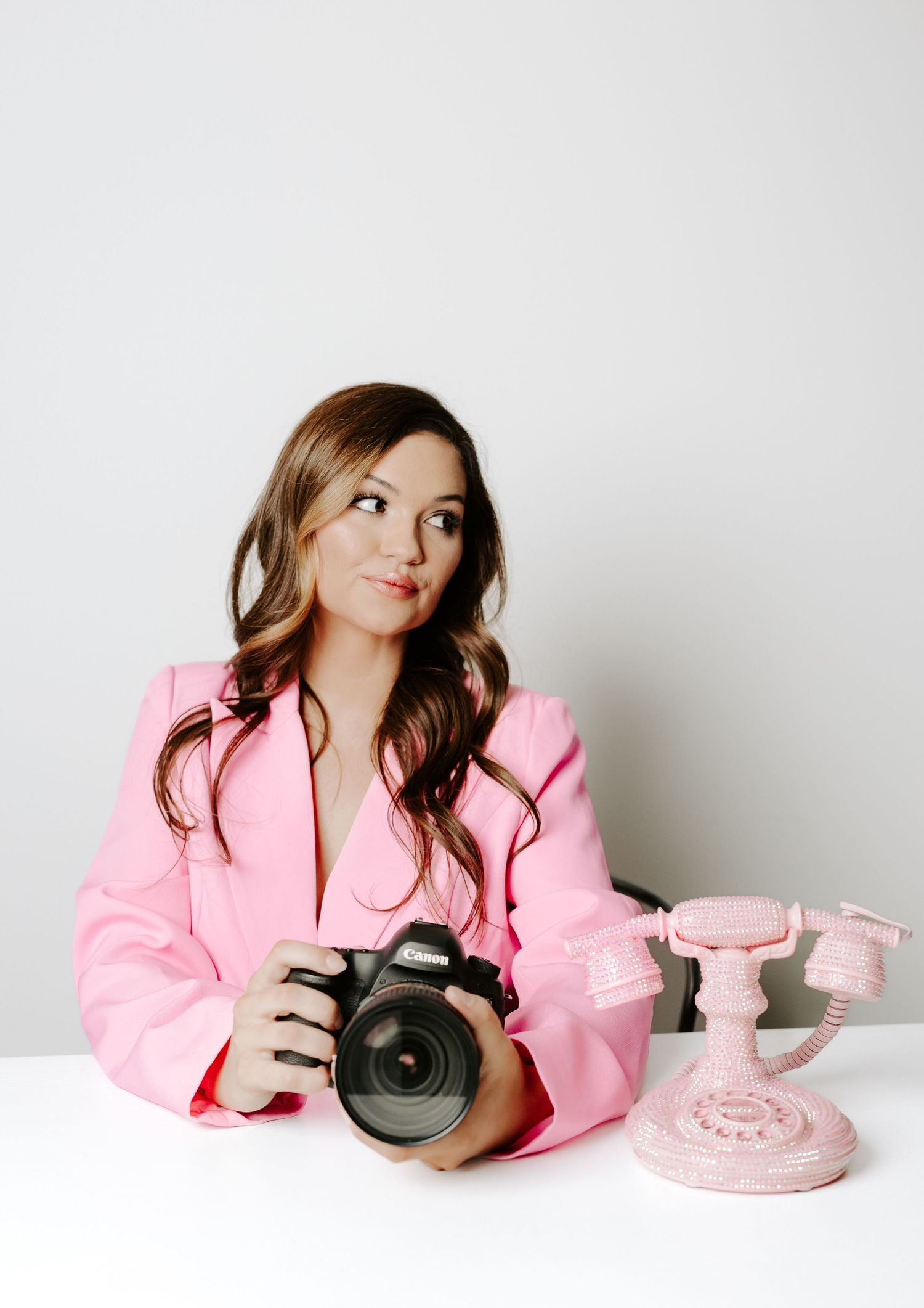 A lifestyle portrait with pink blazer, vintage telephone, and professional camera against white background.