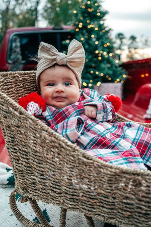 Adorable baby girl in plaid outfit with bow headband smiling in wicker basket at Christmas.