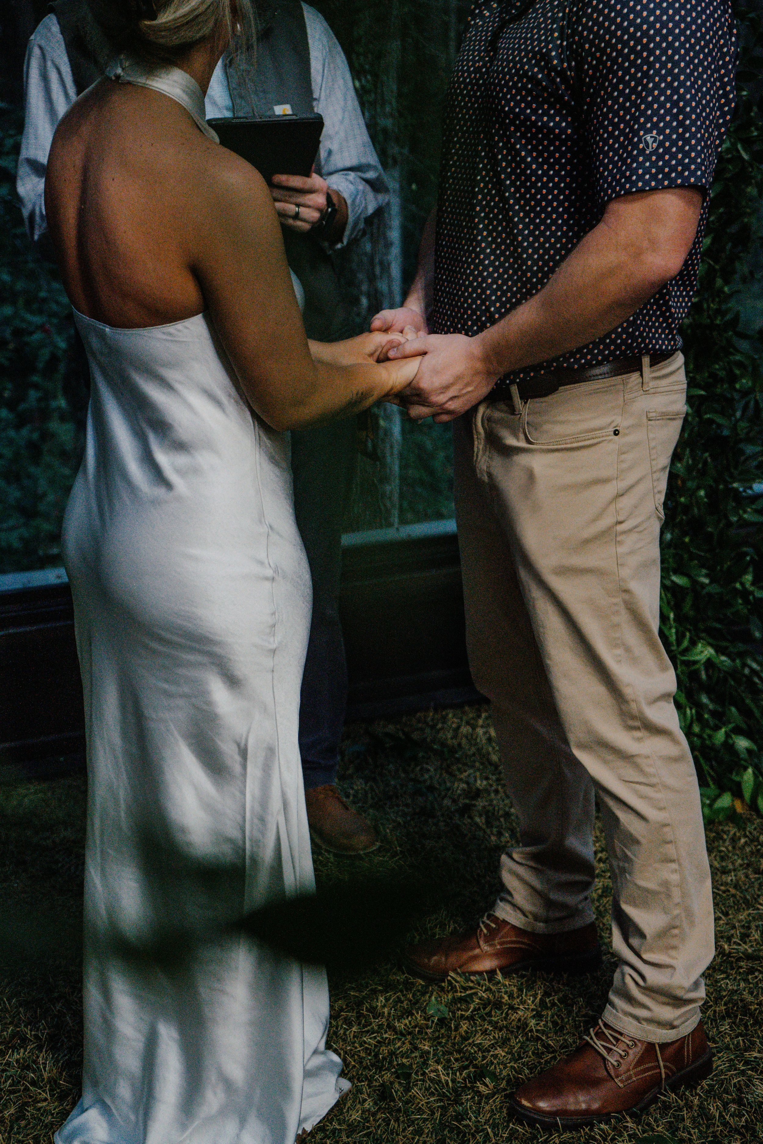 A bride and groom hold hands during an outdoor wedding ceremony before an officiant.