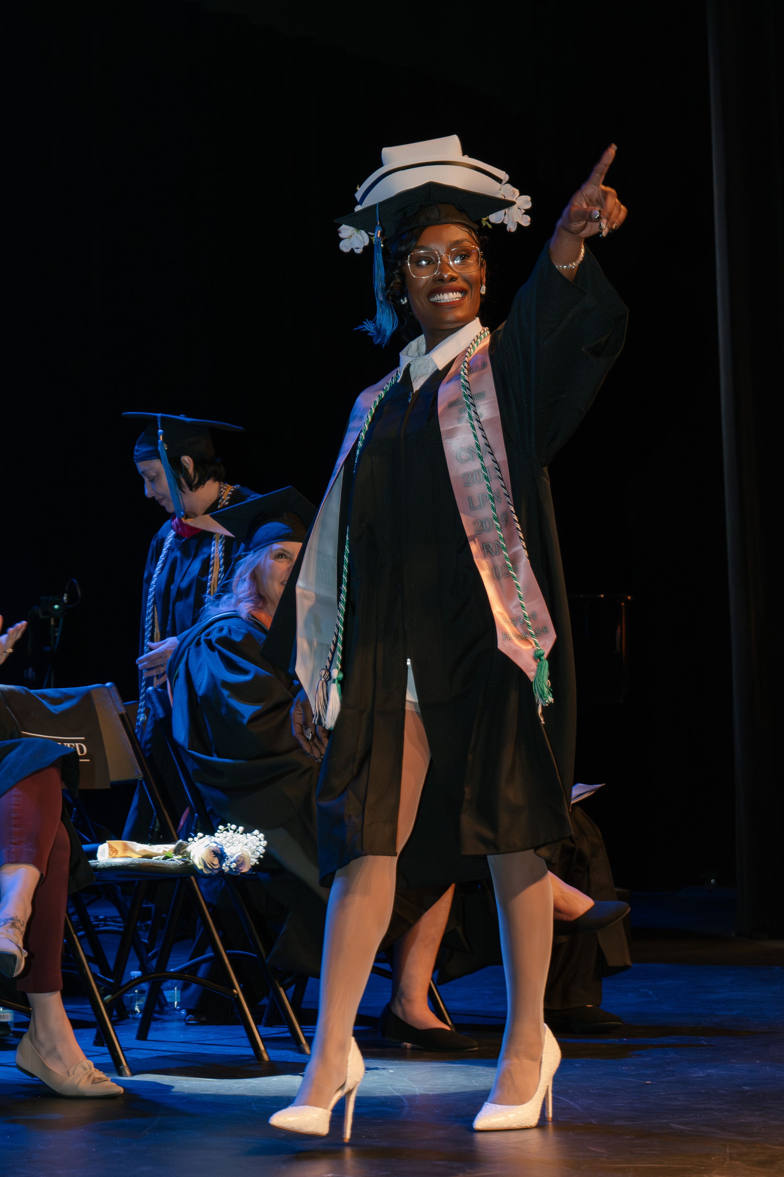 Joyful graduate in black cap and gown pointing upward on stage during graduation ceremony.