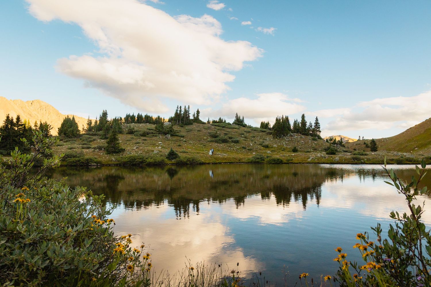 Panoramic mountain landscape with serene alpine lake reflecting clouds and evergreen trees in the wilderness.