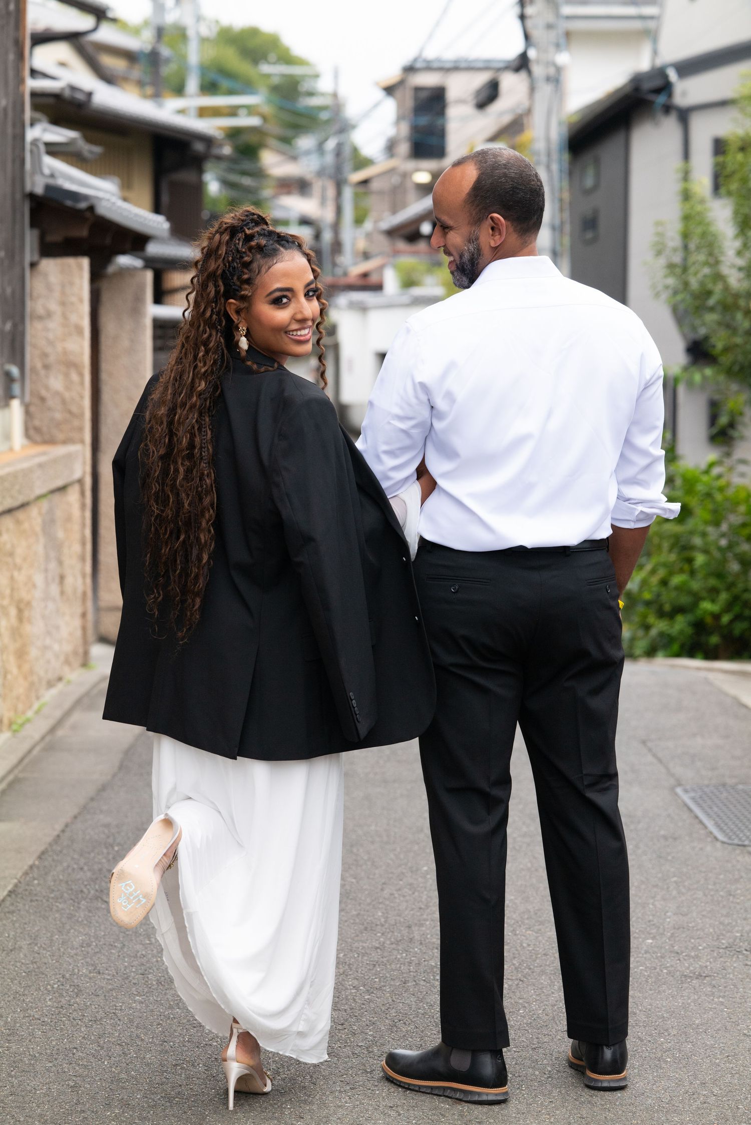 Woman in white dress and black blazer glances back smiling while walking with man on a quiet Kyoto street.