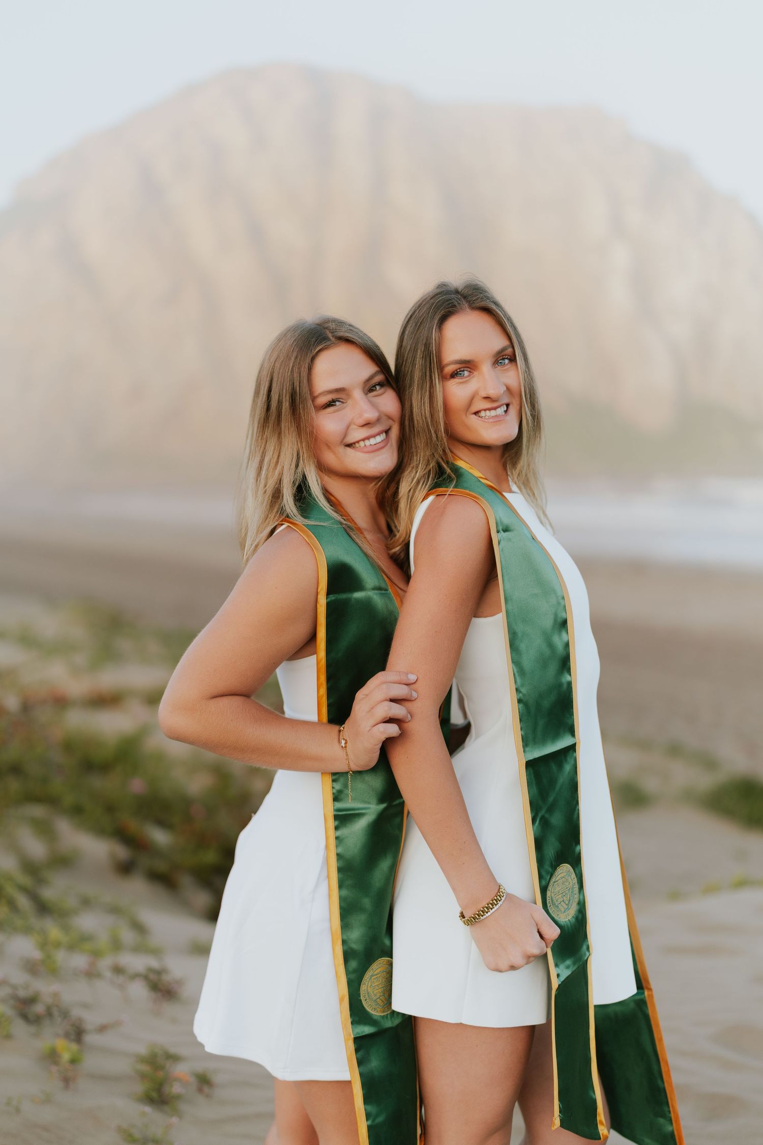 Two graduates in green sashes and white dresses share joyful moments at a beach with mountains in the background.