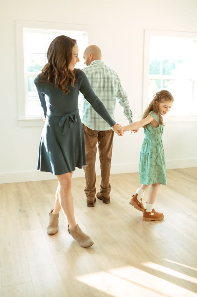 A family dances together in a bright sunlit room with hardwood floors.