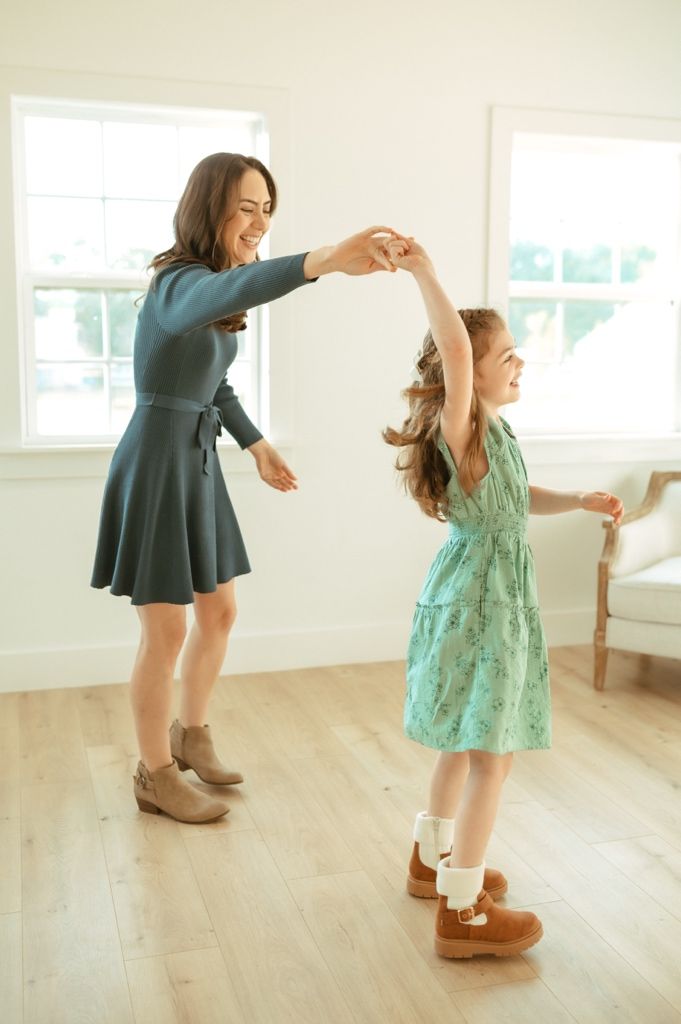 Two people dance together in a bright room, one wearing a teal dress and the other in a mint green dress.