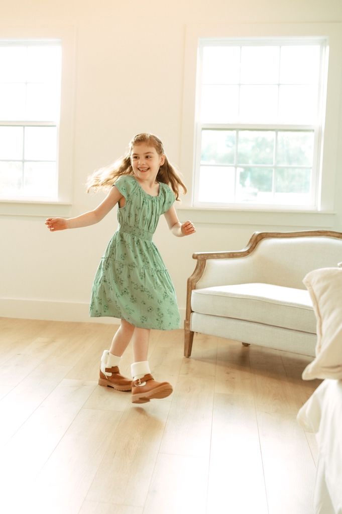 A child in a mint green dress twirls and dances in a bright white room with hardwood floors and elegant furniture.