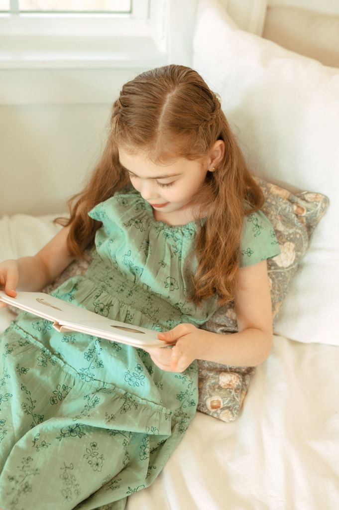 Child in mint green dress reads quietly while resting on decorative pillows near a window.