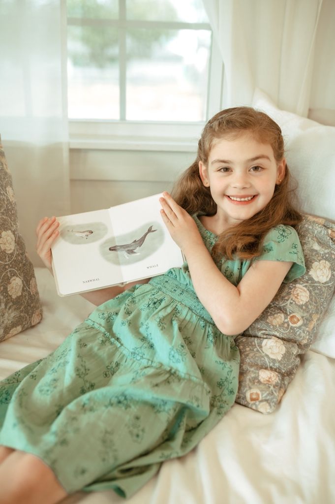 Child in mint floral dress relaxes on cushions while holding a book with a joyful expression.