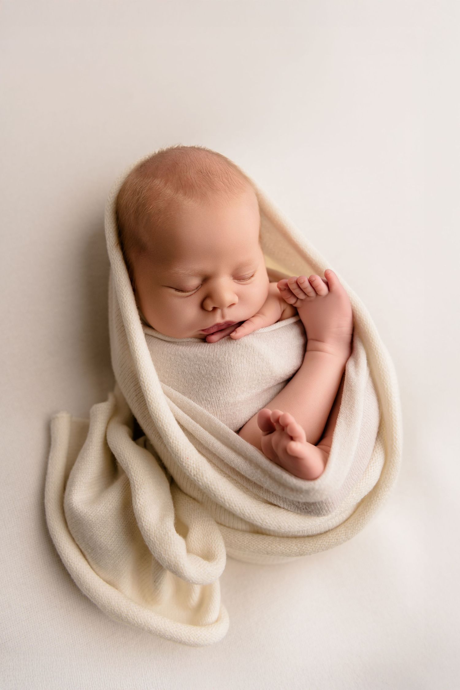 A sleeping baby wrapped in soft beige fabric lies peacefully against a neutral backdrop in a professional portrait setting.