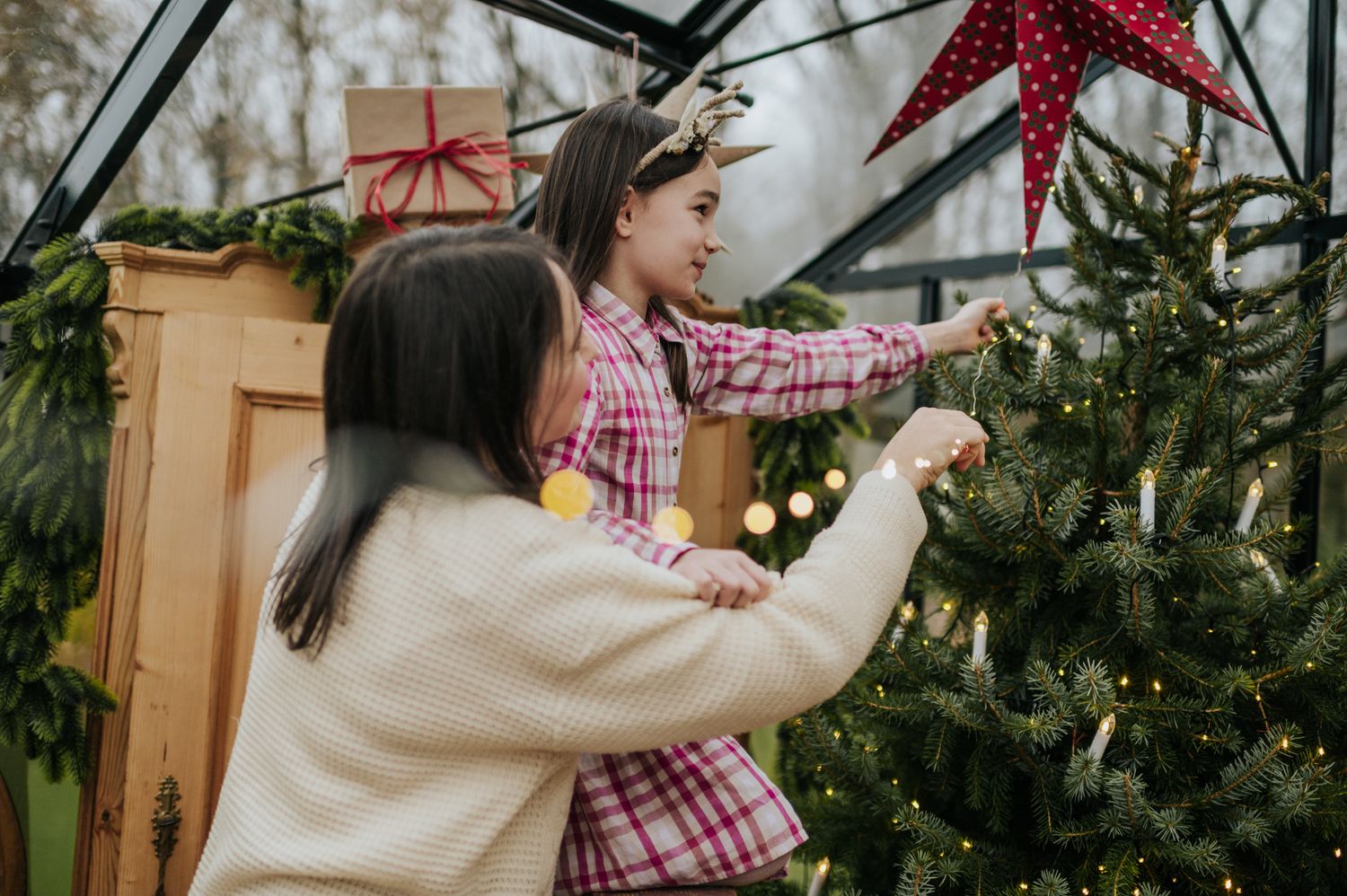 A person in a beige sweater decorates a Christmas tree with red ornaments and string lights in a cozy indoor setting.