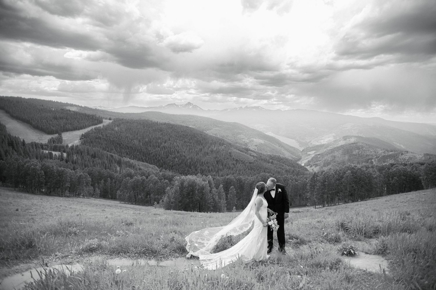 A black and white wedding photo of a couple standing on a hillside with mountain views.
