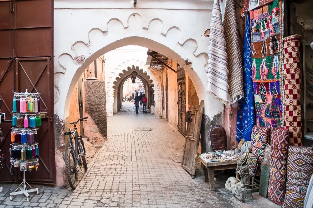 A white arched walkway in a Moroccan market, with textiles and goods displayed along the walls.