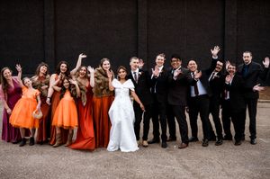 Wedding party group poses together outdoors in formal attire with bridesmaids in orange dresses.