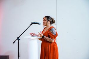 A person in a flowing orange dress records vocals at a microphone in a bright white studio setting.